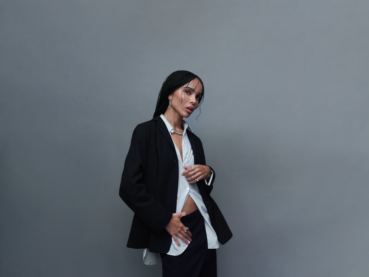 Woman wearing a black blazer and white shirt with a necklace, earrings, and rings, posed against a plain grey background.