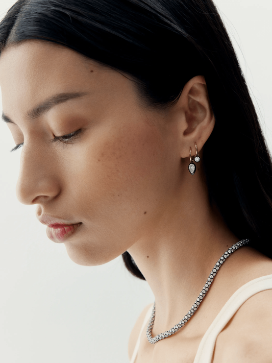 Close-up of a woman wearing pear-shaped diamond hoop earrings crafted in blackened white gold, paired with a diamond tennis necklace, showcasing contemporary and antique-inspired design. View 3