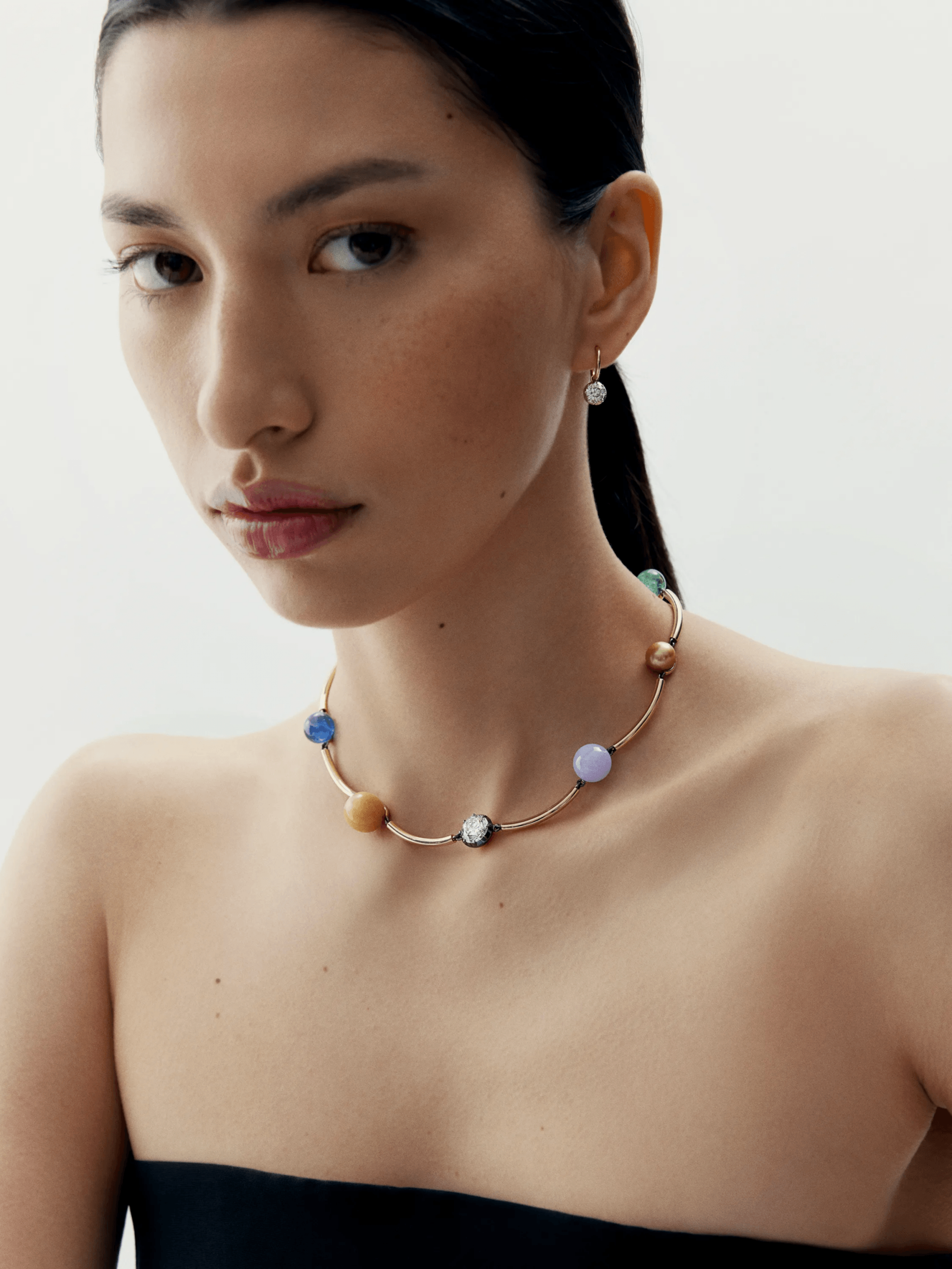 Close-up portrait of a woman wearing a contemporary necklace featuring multicolored round stones set on a sleek gold band, paired with a single diamond drop earring. View 3