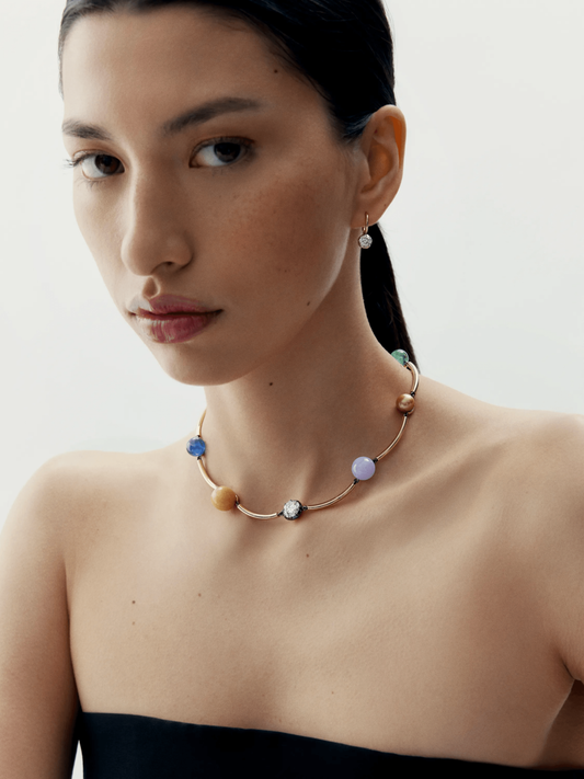 Close-up portrait of a woman wearing a contemporary necklace featuring multicolored round stones set on a sleek gold band, paired with a single diamond drop earring. View 3