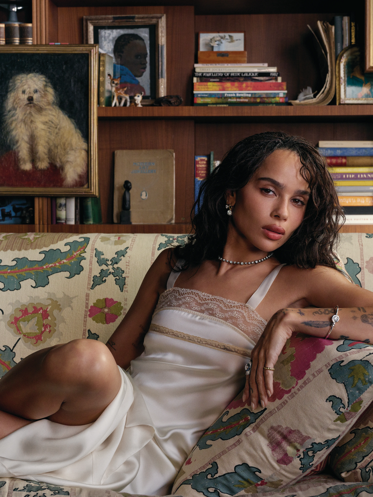 Woman reclining on a patterned sofa in a white satin dress, wearing a delicate diamond necklace, earrings, rings, and a bracelet in a vintage-inspired interior