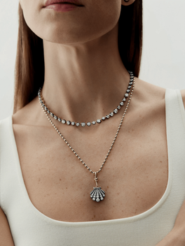Close-up of a woman wearing two layered necklaces, one featuring round diamonds set closely together and the other a detachable pendant shaped like a shell with diamond accents on a ball chain, showcasing handmade craftsmanship with antique-inspired design.