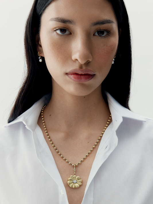 Close-up of a woman wearing a medallion-style flower pendant necklace in warm gold tones with a diamond center, paired with simple hoop earrings, styled with a white shirt. View 3