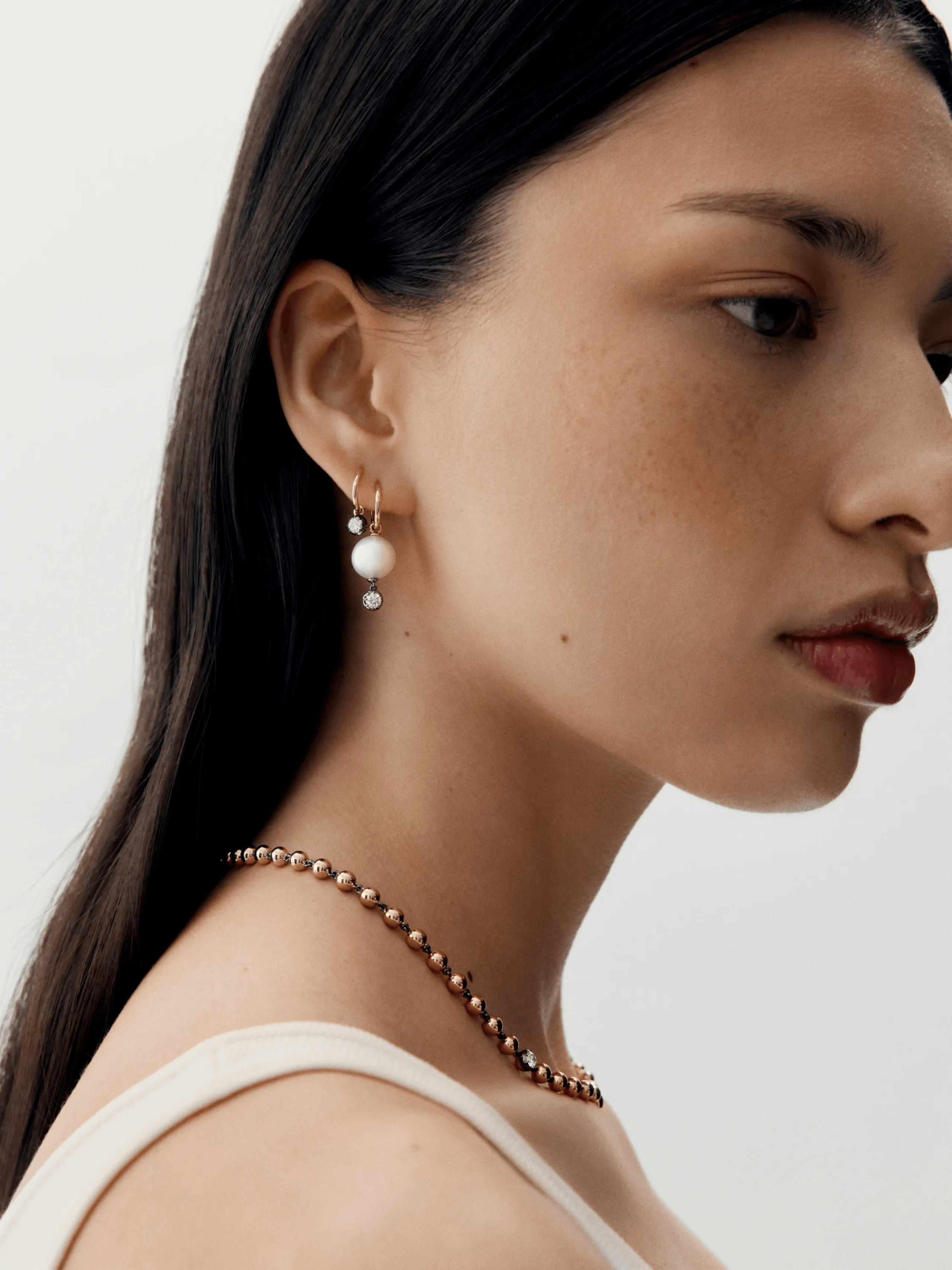 Close-up of a woman wearing delicate gold hoop earrings featuring a pearl and small diamond, paired with a gold beaded necklace, showcasing a minimalist contemporary style. View 3