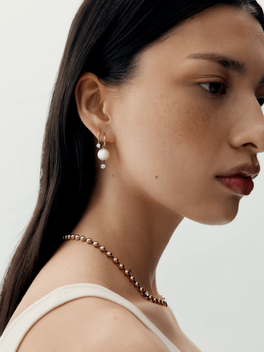 Close-up of a woman wearing delicate gold hoop earrings featuring a pearl and small diamond, paired with a gold beaded necklace, showcasing a minimalist contemporary style.