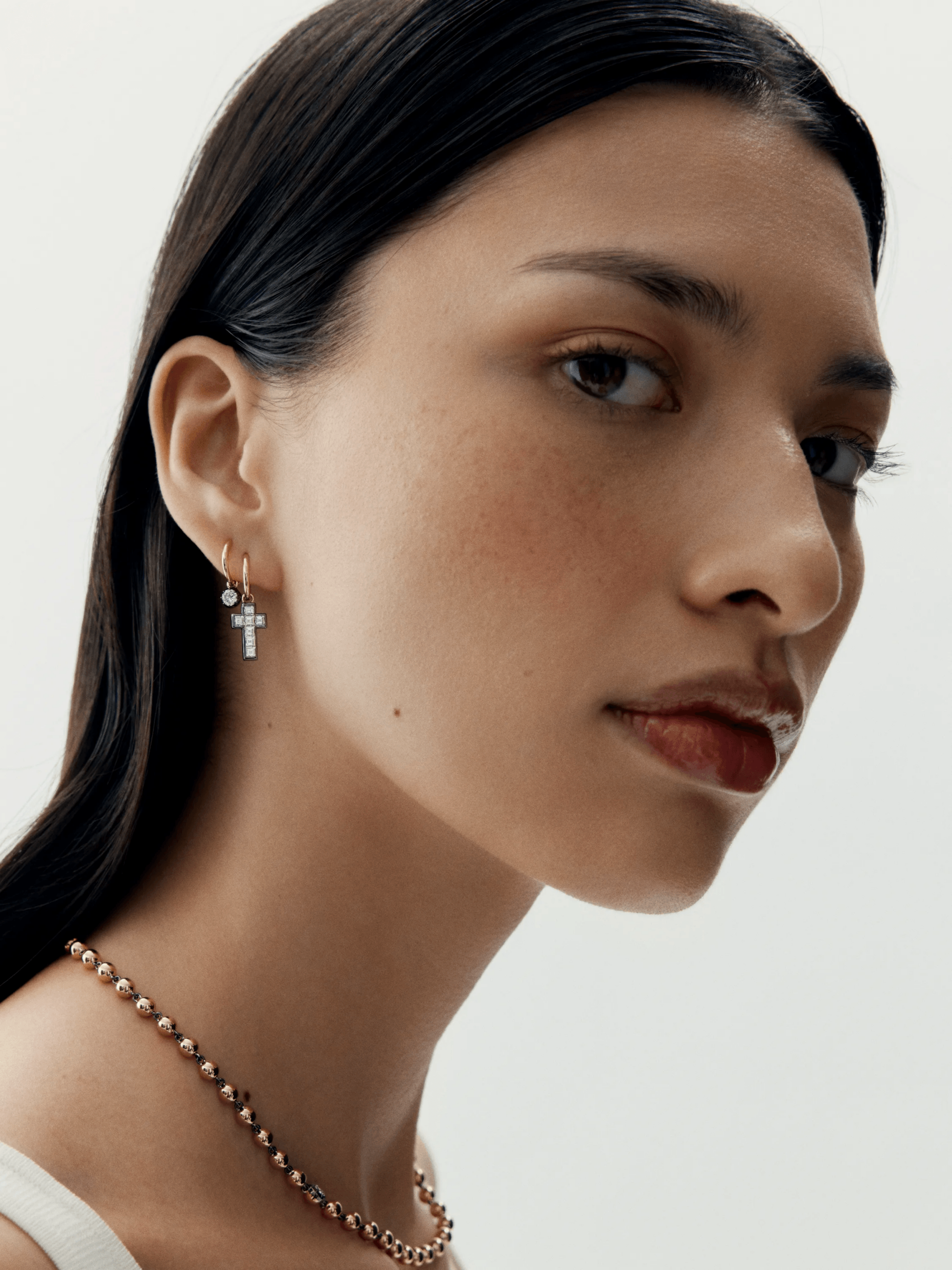 Close-up of a woman wearing a delicate 18K rose gold hoop earring with a carré cut diamond cross and a small round diamond, paired with a beaded necklace, showcasing refined contemporary style and expert craftsmanship. View 3