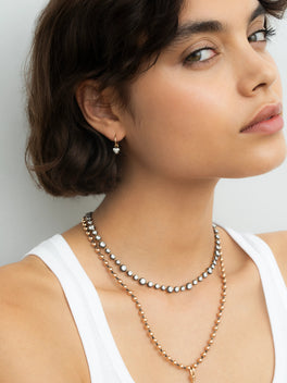Close-up of a woman wearing a delicate blackened gold hoop earring with a small heart-shaped diamond, paired with layered contemporary necklaces in contrasting metals.
