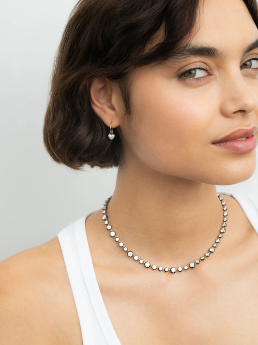 Close-up of a woman wearing a delicate heart-shaped diamond hoop earring in blackened gold, paired with a beaded diamond necklace, showcasing contemporary and antique-inspired craftsmanship. View 3