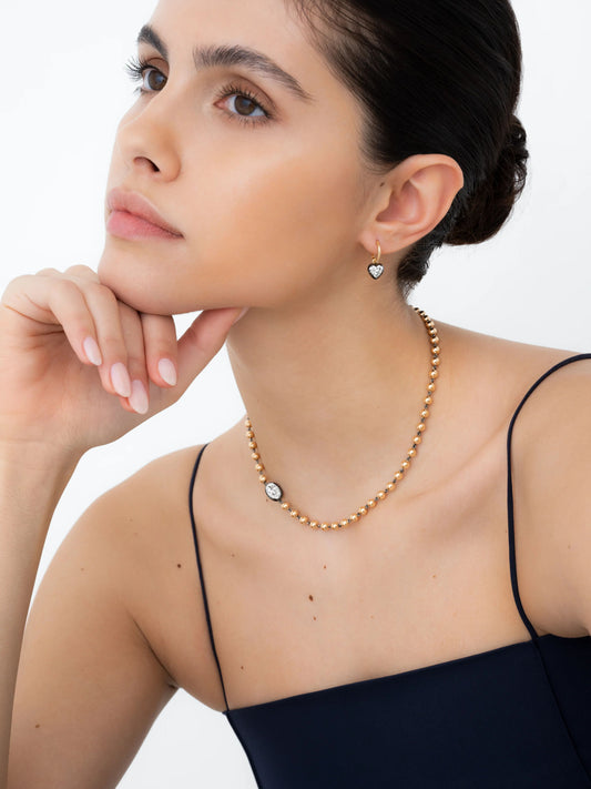 Close-up of a woman wearing a blackened gold hoop earring with a heart-shaped diamond, paired with a delicate gold bead necklace featuring a single prominent round diamond. View 3