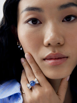 Close-up portrait of a woman wearing a diamond and blue sapphire bow ring, featuring a vivid central sapphire and sculptural diamond ribbons.