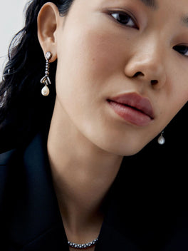 Close-up portrait of a model wearing asymmetric diamond and pearl drop earrings, featuring delicate bow details and luminous pearl pendants in a contemporary style