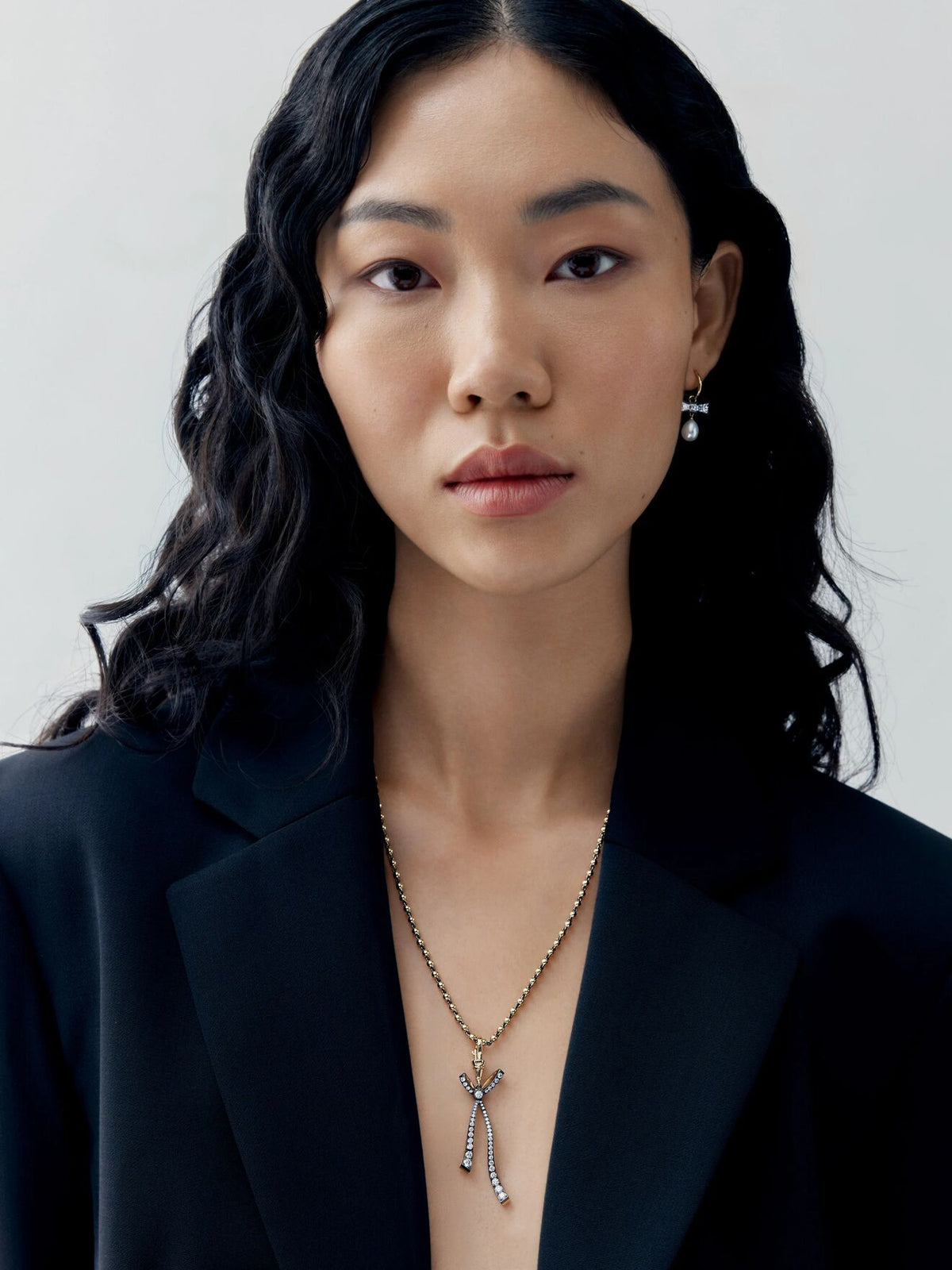 Model wearing a diamond bow pendant with delicate ribbon details and slender drops on a fine ball chain necklace over a black blazer View 3
