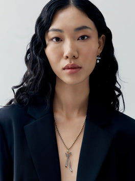 Model wearing a diamond bow pendant with delicate ribbon details and slender drops on a fine ball chain necklace over a black blazer