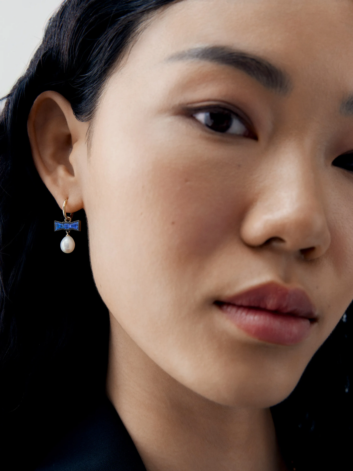 Close-up portrait of a woman wearing a sapphire bow hoop earring with a luminous pearl drop, showcasing Jessica McCormack contemporary jewellery craftsmanship View 3