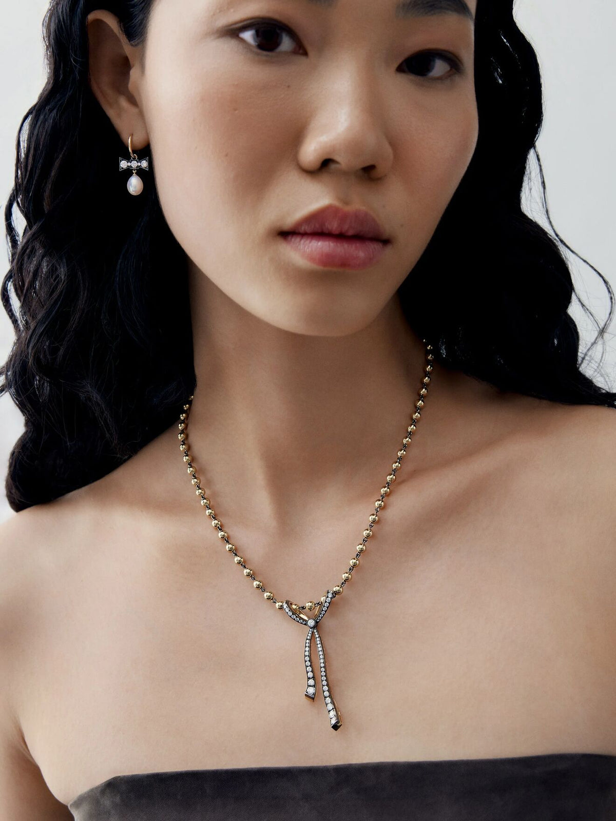 Close-up portrait of a woman wearing a diamond and ribbon pendant necklace on a delicate beaded chain, with slender drop details and a pearl earring. View 3