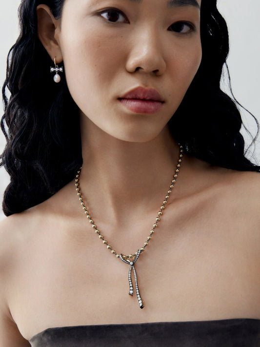 Close-up portrait of a woman wearing a diamond and ribbon pendant necklace on a delicate beaded chain, with slender drop details and a pearl earring. View 3