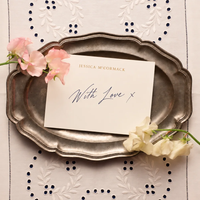 Jessica McCormack handwritten With Love card resting on an antique silver tray, surrounded by pink and white flowers on an embroidered cloth.
