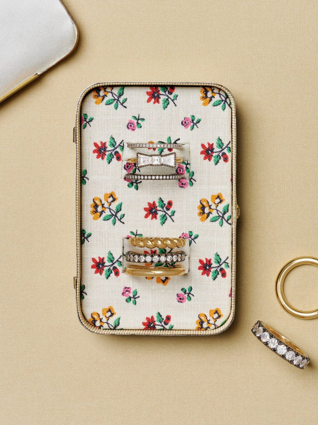 Floral embroidered jewellery case holding stacked rings, with a gold hoop and a diamond set band arranged beside it on a neutral surface