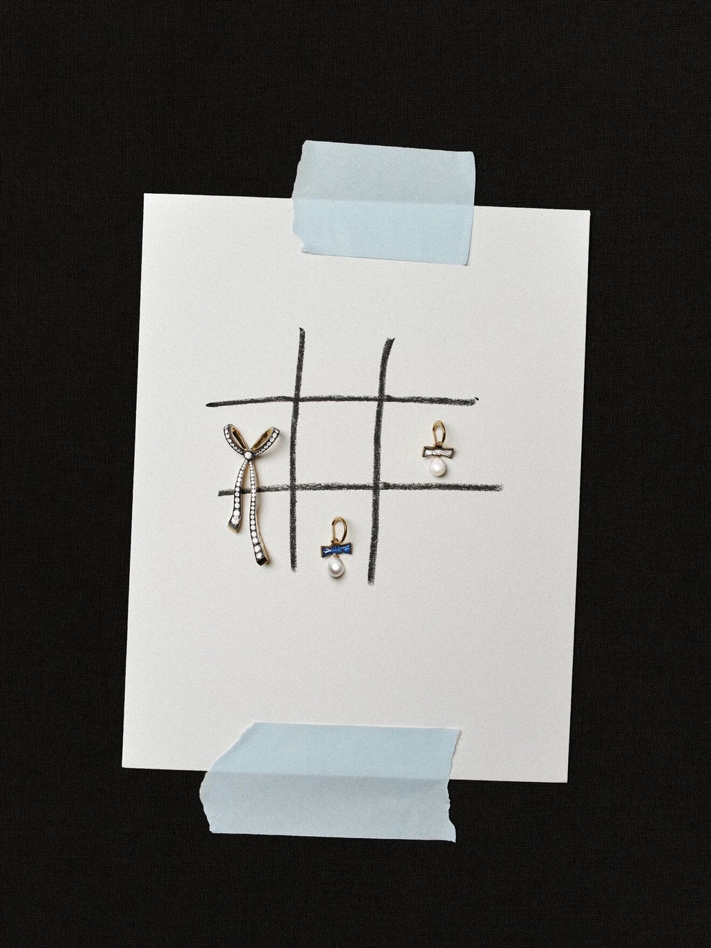 Three delicate earrings arranged on a hand drawn grid on white paper taped against a black background, including a dangling bow shaped earring and pearl details