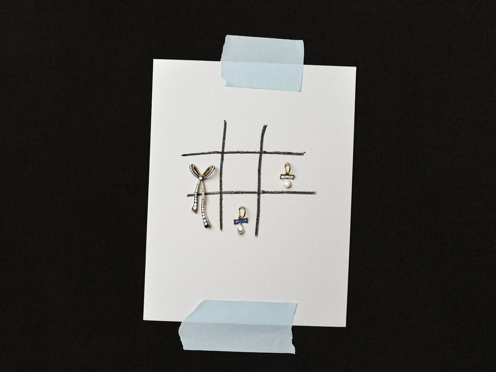 White card taped against a black background, featuring hand-drawn grid lines and three delicate pieces of jewellery arranged across the surface.