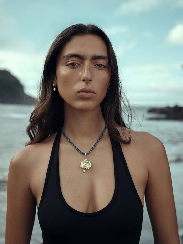 Woman wearing a black top and pendant necklace, standing beside the sea with rocky cliffs and a cloudy sky in the background