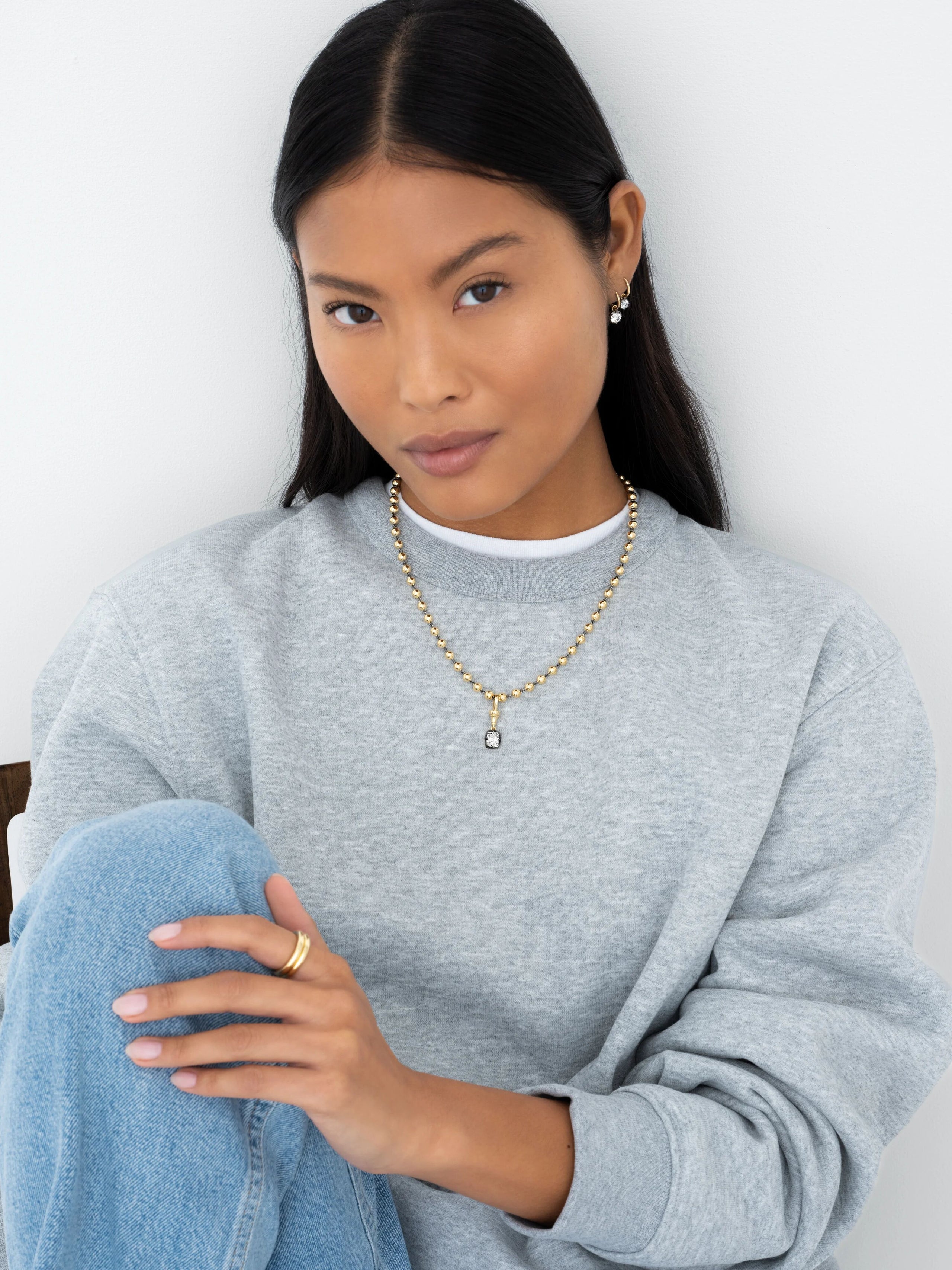 Close-up of a woman wearing a yellow gold beaded necklace with a diamond pendant, styled with casual grey sweatshirt and light blue jeans, showcasing contemporary craftsmanship. View 3