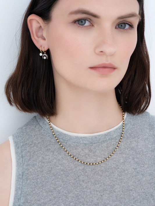 Close-up of a woman wearing a delicate 18-inch yellow gold micro ball chain necklace paired with small gold hoop earrings featuring clear gemstones, showcasing refined, contemporary jewellery. View 3