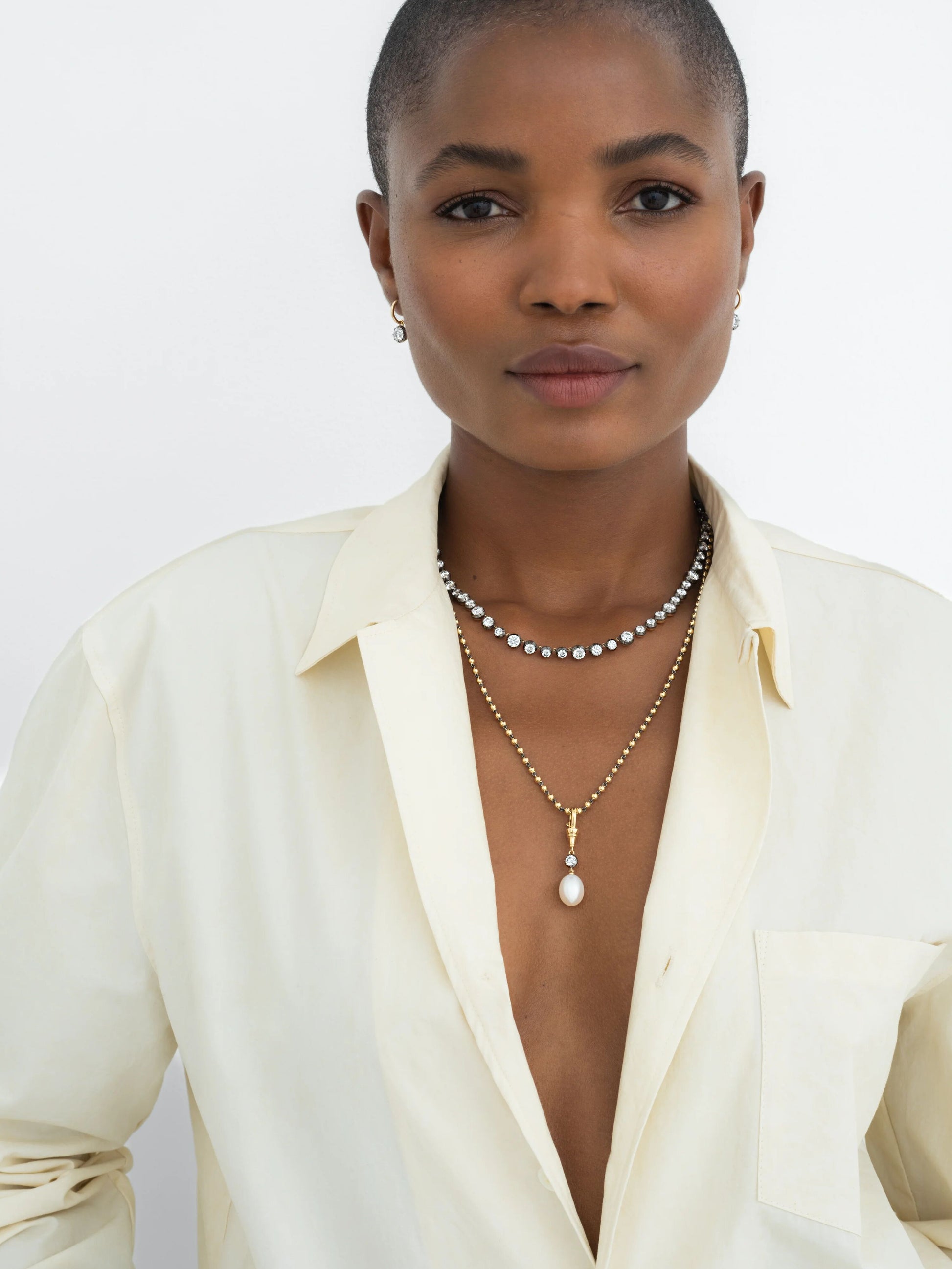 Woman wearing layered necklaces featuring a yellow gold pendant with a pearl and diamonds, paired with a modern diamond necklace, styled with a cream shirt. View 3