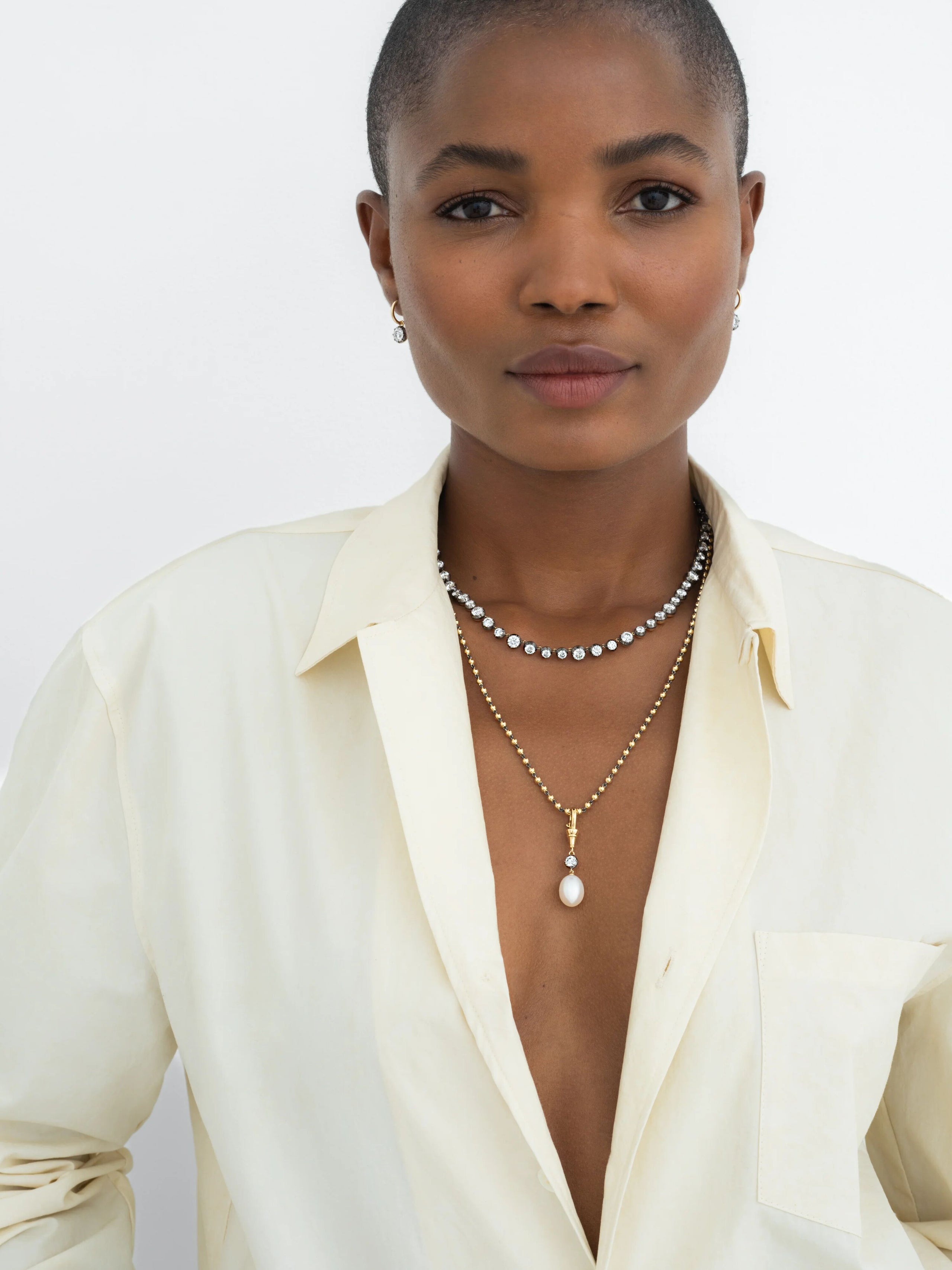 Woman wearing layered necklaces featuring a yellow gold pendant with a pearl and diamonds, paired with a modern diamond necklace, styled with a cream shirt. View 3