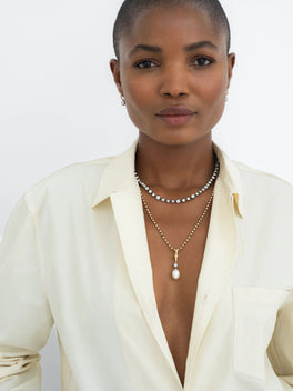 Woman wearing layered necklaces featuring a yellow gold pendant with a pearl and diamonds, paired with a modern diamond necklace, styled with a cream shirt.