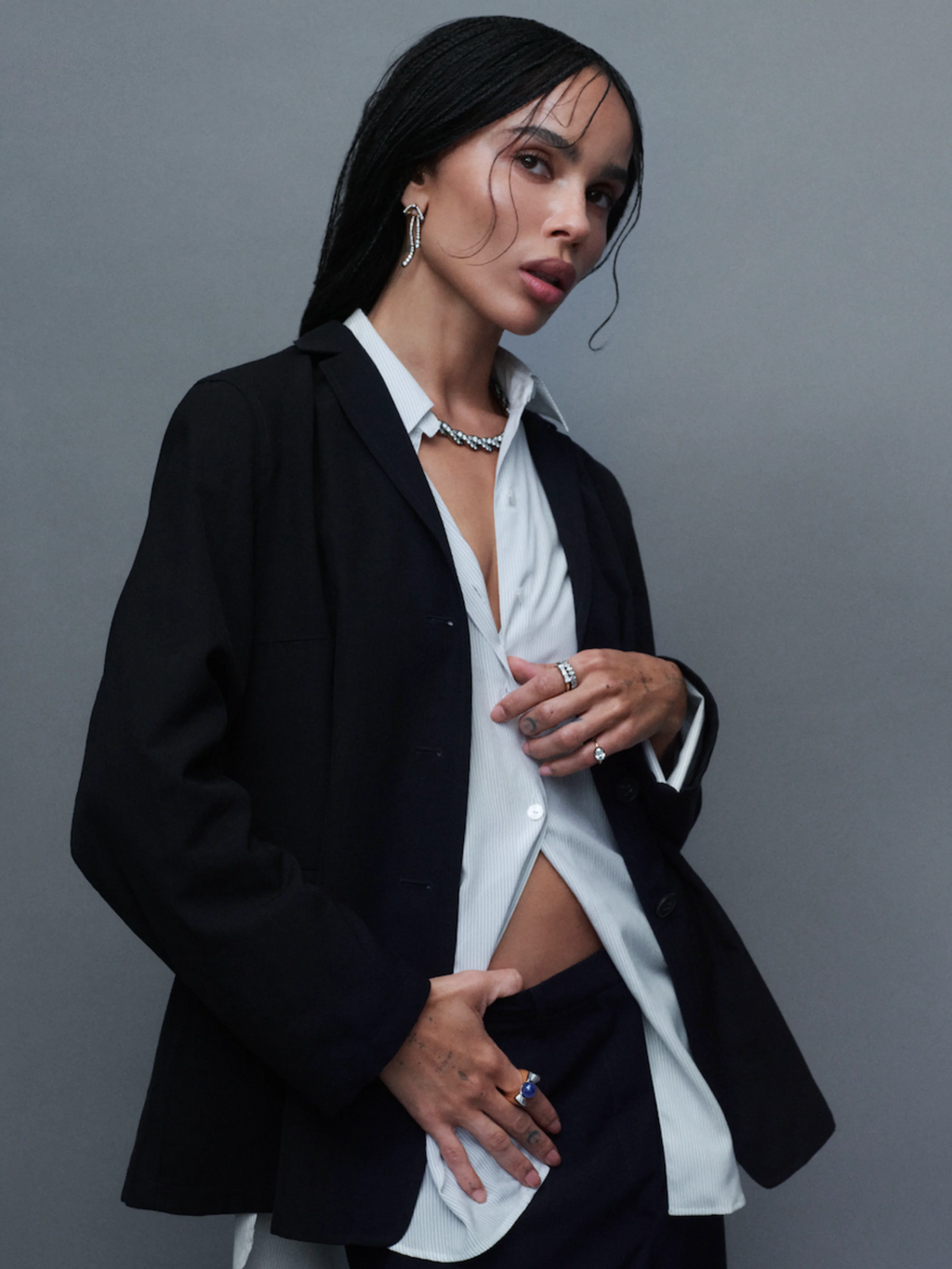 Portrait of a woman in a black blazer and white shirt wearing a necklace, hoop earrings, and several rings against a grey background