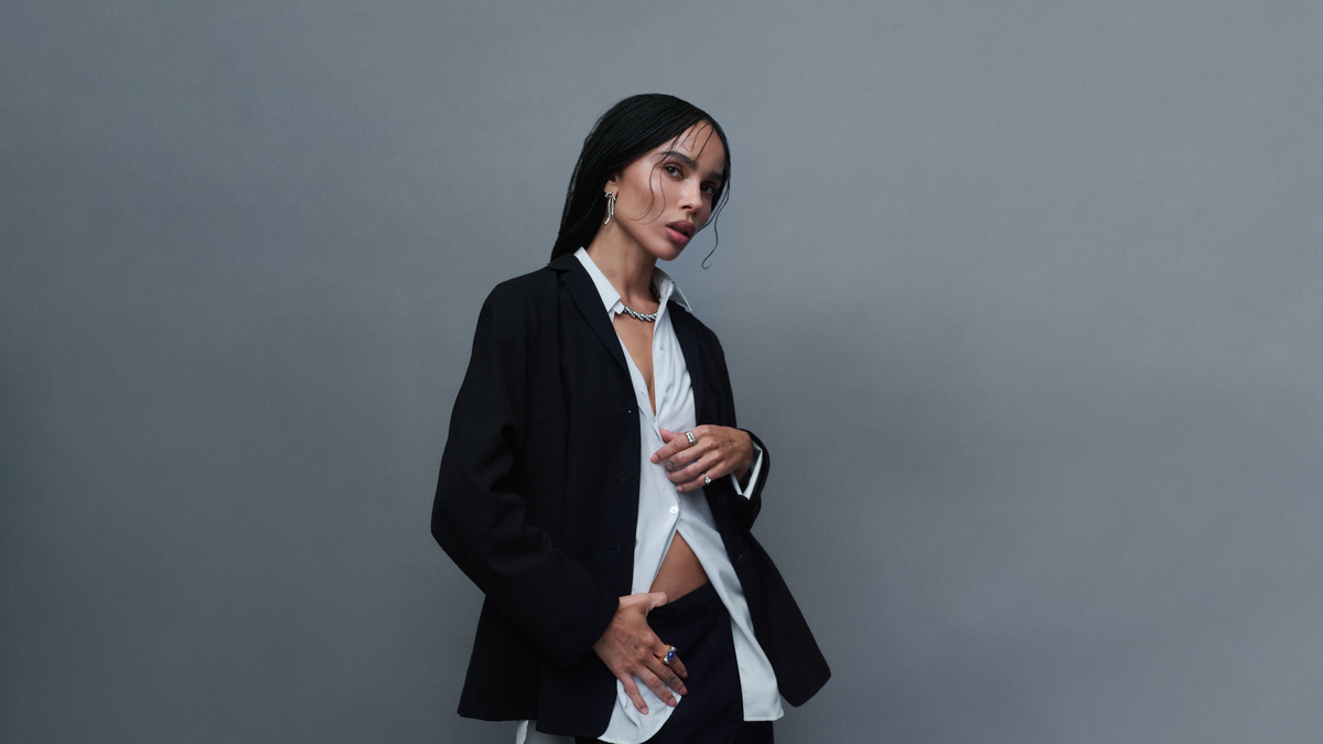 Woman wearing a black blazer and white shirt with a delicate necklace and rings against a plain grey background
