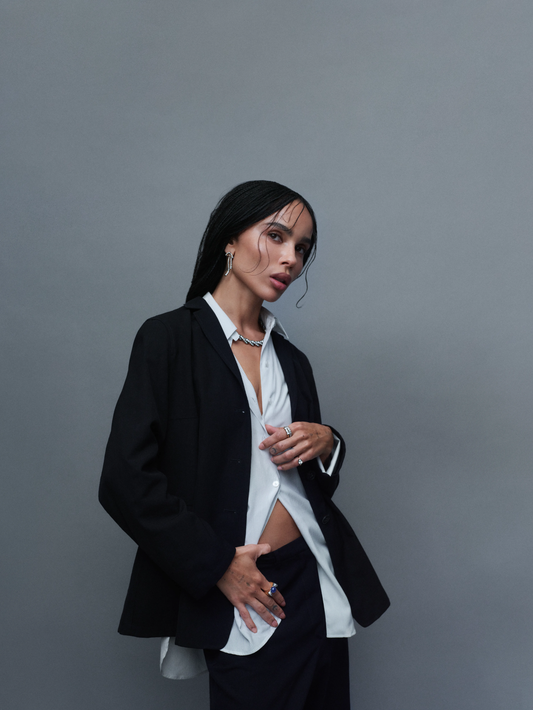 Woman in a black suit and open white shirt wearing a necklace, earrings, and rings against a plain grey background.